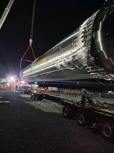 Stainless-clad process vessel lying on its transport trailer with lifting rigging attached under jobsite lighting