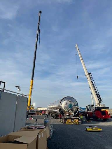 Two all-terrain cranes rigged on either side of a stainless-clad vertical vessel staged on its transport trailer