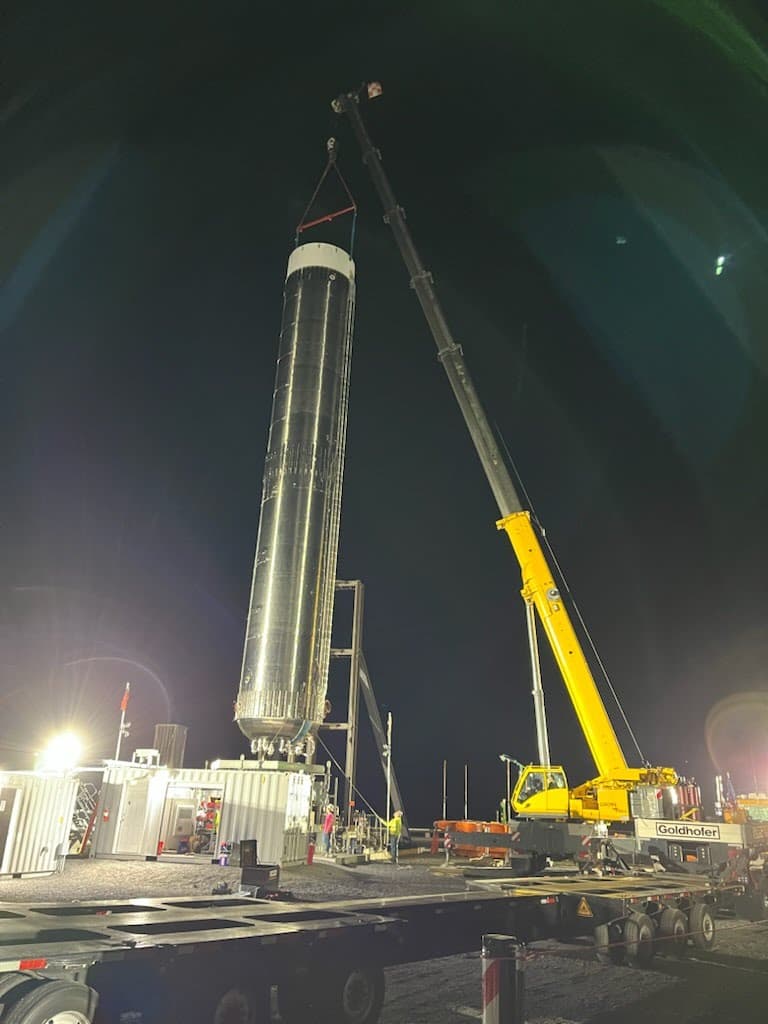 All-terrain crane holding a tall stainless-clad column upright above its foundation while the crew guides it in